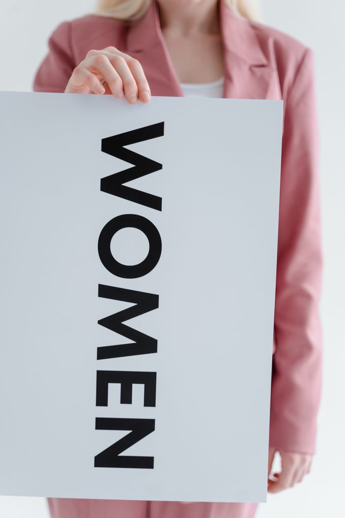 A person in a pink blazer holding a sign with the word 'women' in a studio setting.